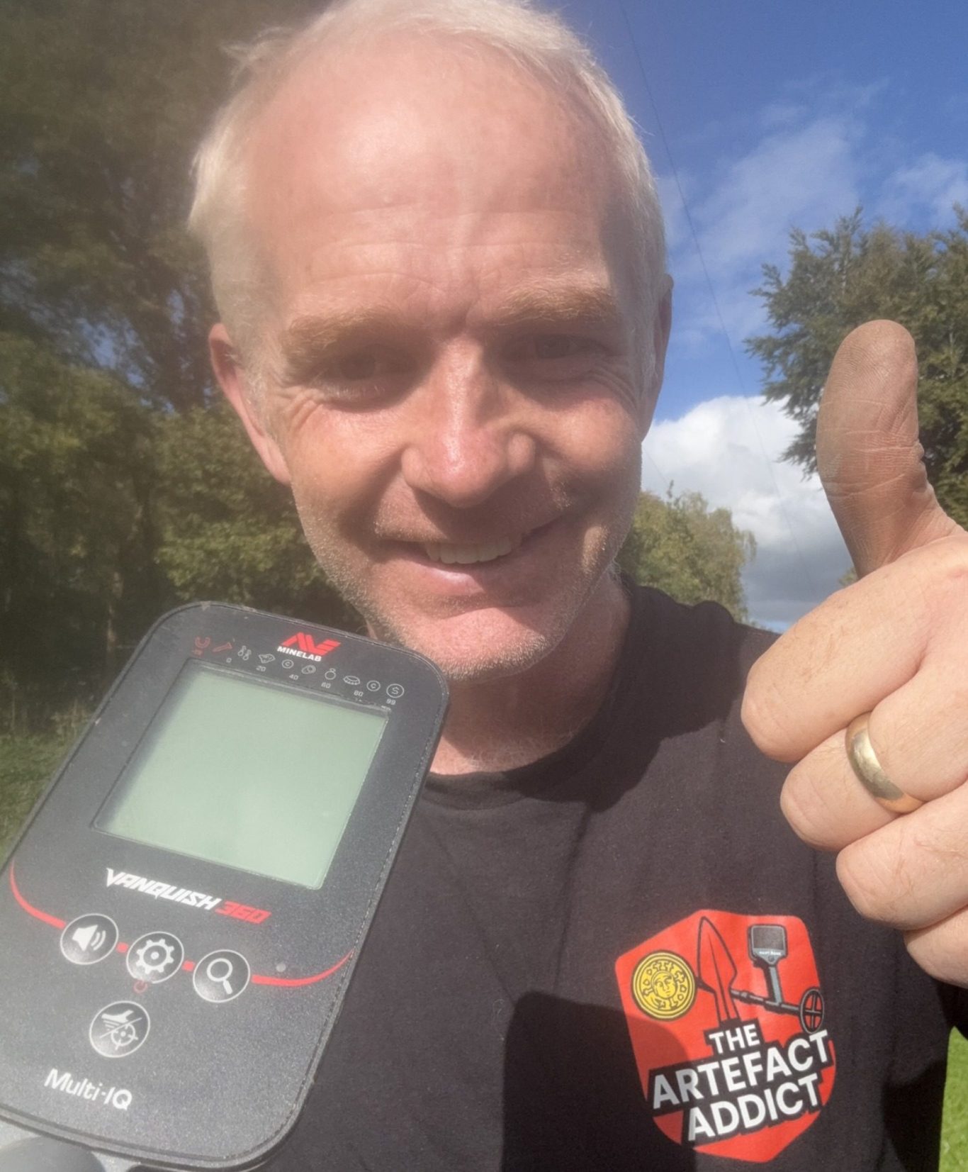 The artefact addict Dave, smiling and giving a thumbs up, holding a metal detector outdoors.