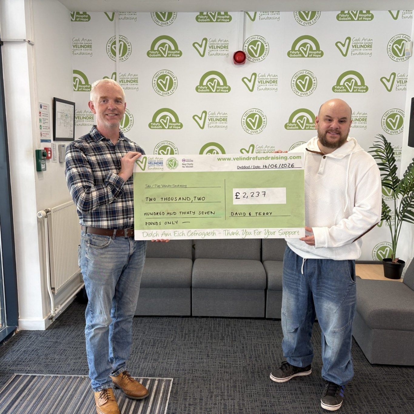 Dave and terry hold a large cheque of £2.237 in a modern office setting with greenery in the background.